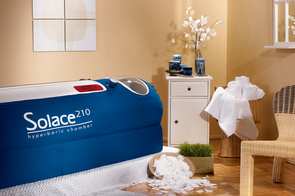 A mild hyperbaric oxygen chamber in vibrant blue, with a cylindrical inflatable bladder and rigid frame. It has three large clear viewing windows set longitudinally, a circular front access port, dual control valves, and visible pressure regulators. The design is compact and the material appears durable and medical-grade.