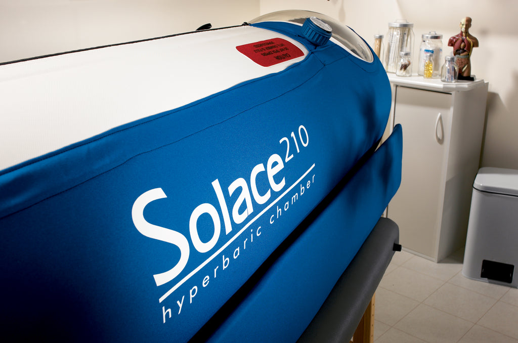 A mild hyperbaric oxygen chamber in vibrant blue, with a cylindrical inflatable bladder and rigid frame. It has three large clear viewing windows set longitudinally, a circular front access port, dual control valves, and visible pressure regulators. The design is compact and the material appears durable and medical-grade.